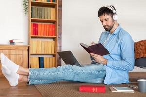 A focused man sits on a rug with a laptop on his lap and a notebook in hand, wearing headphones. A coffee mug, smartphone, and additional books surround him photo