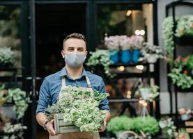 Small business and start of working day. Man in protective mask takes out box of plants outside in front of flower shop photo