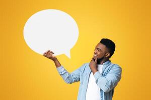Smiling black millennial guy holding empty speech bubble and touching his chin over yellow studio background, copy space photo