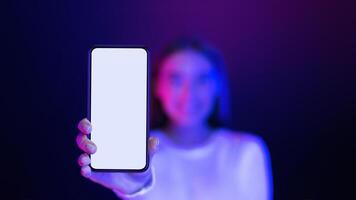 A woman is standing in front of a purple background, holding up a phone with a blank screen. She appears to be showcasing the device, mockup, copy space photo