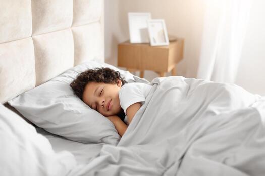 un joven africano americano chico, en pijama, pacíficamente duerme en un acogedor cama cubierto con un blanco edredón. el habitación es tenuemente iluminado, y el niño es rizado arriba cómodamente, descansando pacíficamente foto