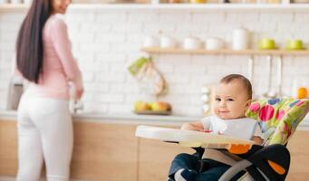 A smiling baby sits comfortably in a colorful high chair in a modern kitchen while the mother is busy in the background preparing food. photo