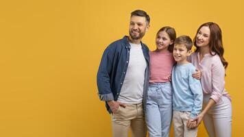 A happy family, with two parents and two children, embrace each other against a vibrant yellow backdrop. They appear cheerful and content, looking at copy space photo