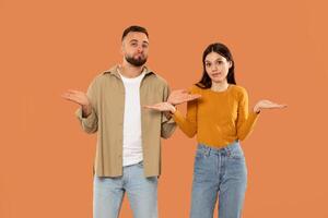 A man and woman stand side by side, both raising their hands in a gesture of confusion. They are dressed casually, orange studio background photo