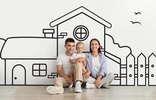 A young couple is sitting on the floor holding their toddler, smiling at the camera with a large hand-drawn illustration of a house and surroundings on the wall behind them photo
