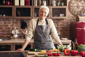 A smiling senior woman in an apron stands in a modern kitchen with exposed brick and a red refrigerator. She has prepared a fresh salad with cucumbers, peppers, and cherry tomatoes. photo