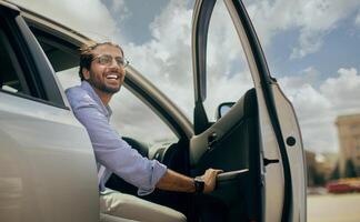Positive young middle-eastern man going out the car, low angle photo, copy space. Happy arab guy open automobile door and smiling, getting in. Auto test-drive, leasing, buying, renting concept photo