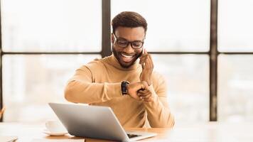 Punctuality Concept. Happy black man checking time on wrist watch and talking on mobile phone, empty space, panorama photo