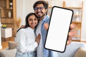 A joyful Indian couple stands in their cozy living room, smiling at the camera while one of them holds up a blank smartphone. They exude happiness and warmth in their casual attire. photo