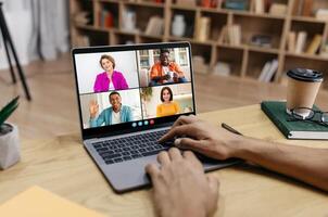 Guy uses a laptop to participate in a call with four other people. The call is taking place in a home office setting, with a bookshelf in the background. photo