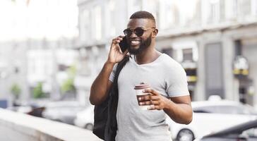 A man in sunglasses smiles while talking on a cell phone and holding a coffee cup in his hand. He is walking down a city street, and cars can be seen in the background. photo