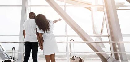 African American couple stands near an airport window, gazing out at the view, while holding hands and waiting for their flight. The woman is pointing at something out of frame, copy space photo