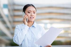 Asian woman stands outdoors, focused on a phone conversation while holding a sheet of documents. She appears to be discussing important matters, with a concerned expression on her face photo