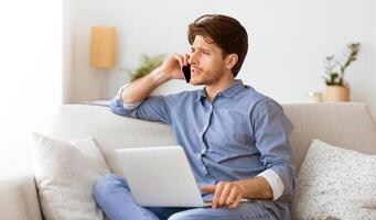 A man sits on a couch, talking on a phone while working on a laptop. photo