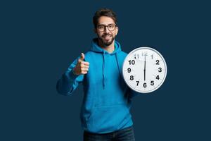 A man wearing a blue hoodie is shown holding a clock in his hands, looking at the time. The mans face is expressionless, and the focus is on the clock he is holding. photo