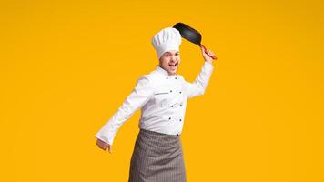 Funny Chef Man Jumping Raising Frying Pan Having Fun Posing Over Yellow Studio Background. Full Length photo
