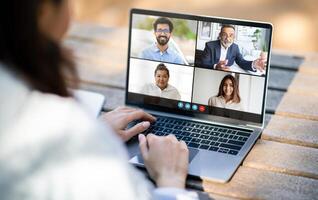 Woman is participating in a virtual meeting on a laptop placed on an outdoor table. Four colleagues are visible on the screen, each engaging in conversation from different locations photo