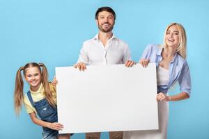A happy family of three is standing in front of a blue background, holding a large, blank sign. They are all looking at the camera. photo