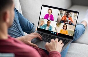 A man sits on a couch with his legs extended and uses a laptop computer to participate in a conference call. The laptop screen displays four different feeds of other people photo