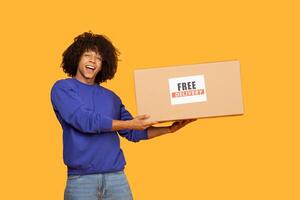 A young man with curly hair smiles broadly while holding a large cardboard box labeled with a free delivery sign. The bright yellow background, the excitement of receiving a package. photo