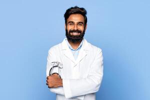 Indian man with a beard, wearing a white lab coat, stands against a light blue background. He is smiling and has his arms crossed, holding a stethoscope. photo