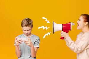 Modern kids problems. Gadget addicted boy playing mobile games on cellphone, ignoring his screaming with megaphone mother, orange background photo
