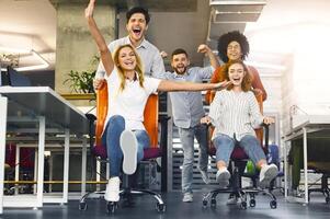 A group of young office workers is caught in a moment of spontaneous joy, racing each other in office chairs. Their expressions are filled with laughter and excitement, suggesting a fun break photo