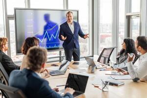 A businessman stands in front of a large screen displaying a financial chart, pointing to the data and presenting it to a group of colleagues in a modern office. photo