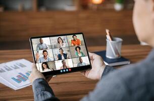 A person is sitting at a desk and looking at a tablet computer. The tablet is displaying a conference with eight people. The people are all smiling and looking at the camera. photo