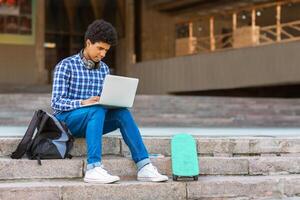A teen black guy with short, curly hair sits on a set of concrete steps in front of a building, using a laptop computer. A black backpack sits beside him, and a teal skateboard is resting on the step photo