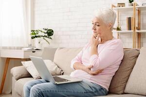 Senior woman is seated on a couch, intently using a laptop computer in a relaxed posture. She appears focused on the screen, with a comfortable expression. photo