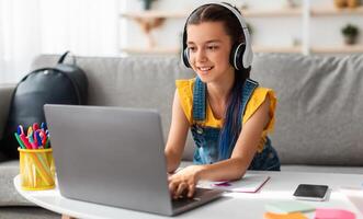 Portrait of little girl in wireless headset using laptop, studying online at home, interested happy student typing on keyboard looking at pc screen, watching webinar, online course, doing homework photo