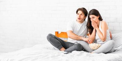 A couple sits on a bed, both looking scared and holding a bowl of popcorn, likely while watching a scary movie. They are both dressed casually in grey pants and white shirts. photo