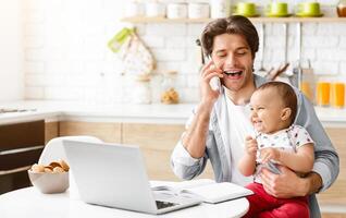 A father is sitting at a kitchen table with his laptop in front of him. He is talking on the phone and smiling while holding his baby. The baby is also smiling and looking at copy space photo