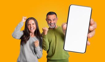 Mature man and woman are standing in front of yellow background, both looking at the camera and smiling excitedly with their fists raised in the air, holding a smartphone with a blank white screen photo