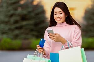 Asian woman is seen holding multiple shopping bags and credit card in one hand while using a cell phone in the other hand. She appears to be multitasking, possibly checking her messages photo