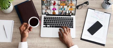 A person is sitting at a desk in an office setting, working on a laptop while participating in a conference call. They are holding a cup of coffee in their left hand, web-banner photo