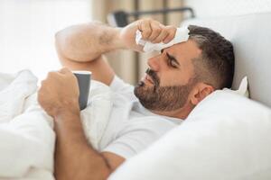 A man lies in bed with a tissue in his hand, wiping his forehead. He appears to be sick or unwell, as he holds a mug and has a tired expression on his face. He is covered by white bedsheets photo