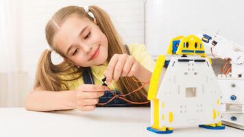 A young girl with pigtails smiles as she builds a robot kit at a white table. She is holding a wire and connecting it to the robot, which is white and yellow. photo