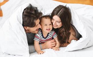 This image shows a happy family of three in their bed. The mother, father, and baby are all smiling and looking at each other. photo