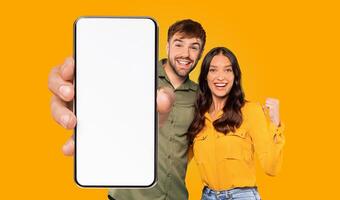A happy couple is holding a smartphone with a blank white screen, and they are both pointing at the screen with smiles on their faces. They are standing in front of a bright yellow background. photo