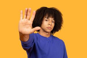 This image shows a young man with curly hair standing in front of a yellow background. He is holding his hand up in a stop gesture. His expression is serious and his eyes are focused on the camera. photo