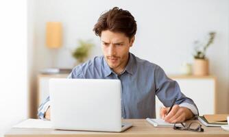 A man sits at his desk in a home office, working on a laptop and taking notes in a notebook. photo