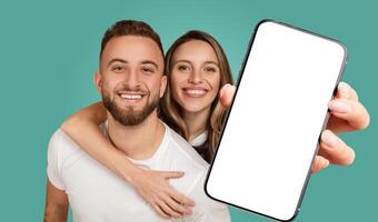 A young couple stand in front of a turquoise background, smiling happily. The woman has her arm around the man's shoulders, and she is holding up a smartphone with a blank screen photo