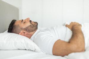 A man with a beard is sleeping on his side in a bed. He is lying on a white pillow, with a white sheet pulled up to his chest. The room is bright and the bed is white. photo