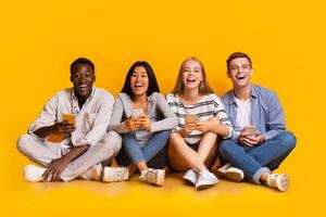 New funny mobile app. Laughing group of multiracial classmates holding smartphones and looking at camera, yellow studio background, panorama photo