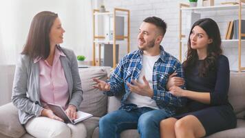 A young couple sits on a couch in a modern office setting, while a therapist listens attentively. The man is speaking with his hands, while the woman sits quietly beside him. photo