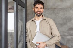 A man with a beard and short brown hair smiles as he holds a laptop computer and leans against a glass door in an office building. The man is wearing a tan button-down shirt and a white undershirt. photo