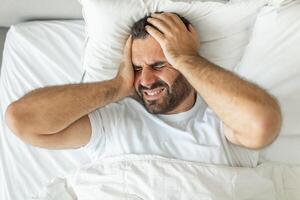 A man lies in bed, his face contorted in pain as he holds his head with both hands. He is likely suffering from a headache or migraine, his expression showing discomfort and distress, top view photo