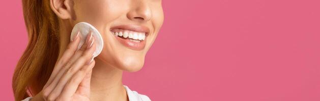 A close-up image of a woman with long brown hair smiling while applying a cotton pad to her cheek in front of a solid pink background. The womans eyes are not visible, cropped, blank space photo